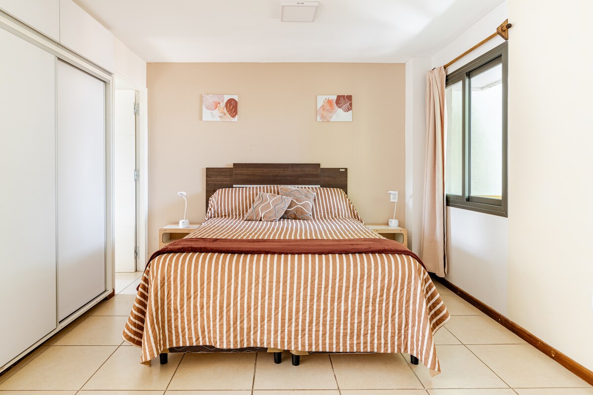 A spacious bedroom features a striped brown and beige blanket atop a neatly made bed. Two decorative paintings adorn the wall above the bed. Natural light filters in through a large window, complemented by neutral-toned walls and tiled flooring.