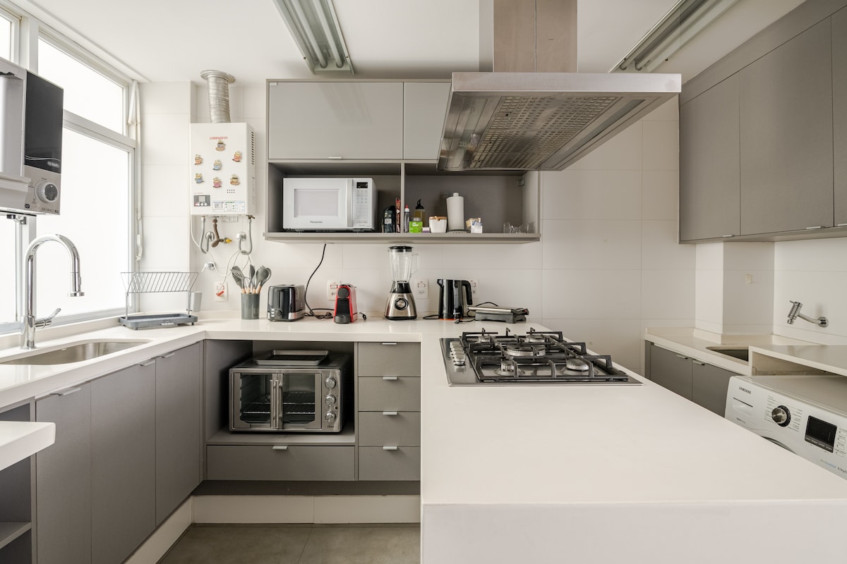 The kitchen features modern cabinetry in neutral tones, equipped with a gas stove, microwave, and various appliances including a toaster and coffee maker. A large island counter provides additional workspace and seating, with ample natural light streaming through large windows.