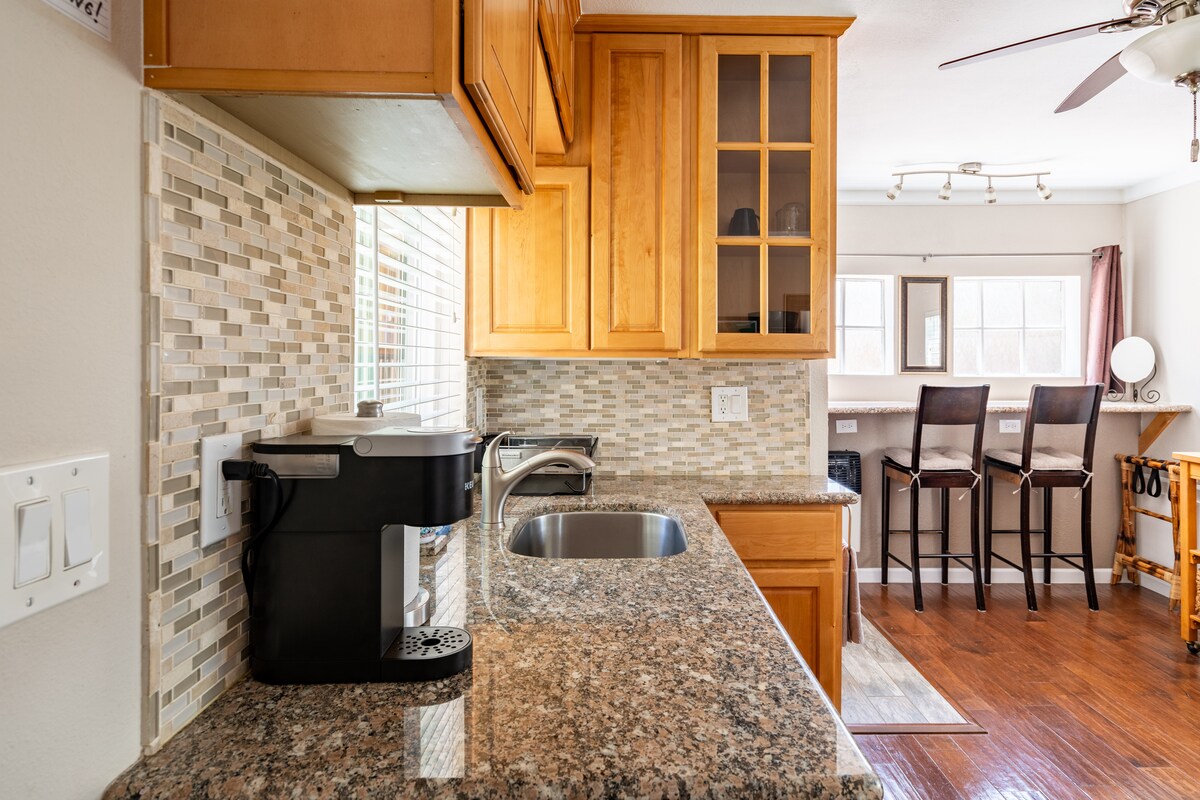 A granite kitchenette is displayed, featuring a coffee maker and sink beneath a window. Wooden cabinets provide storage above, complemented by a bar with high stools in the background. Natural light fills the space, highlighting the neutral tones and textures.