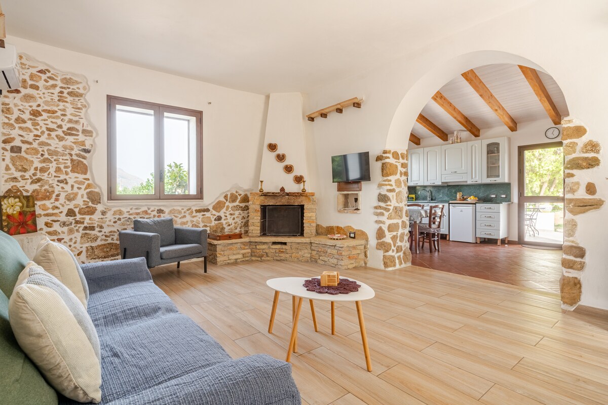 A spacious living area features a stone wall and a cozy fireplace, enhancing the rustic charm. Comfortable seating is arranged around a central coffee table, with soft cushions adding comfort. Natural light streams through the windows, illuminating the adjoining kitchen visible in the background.