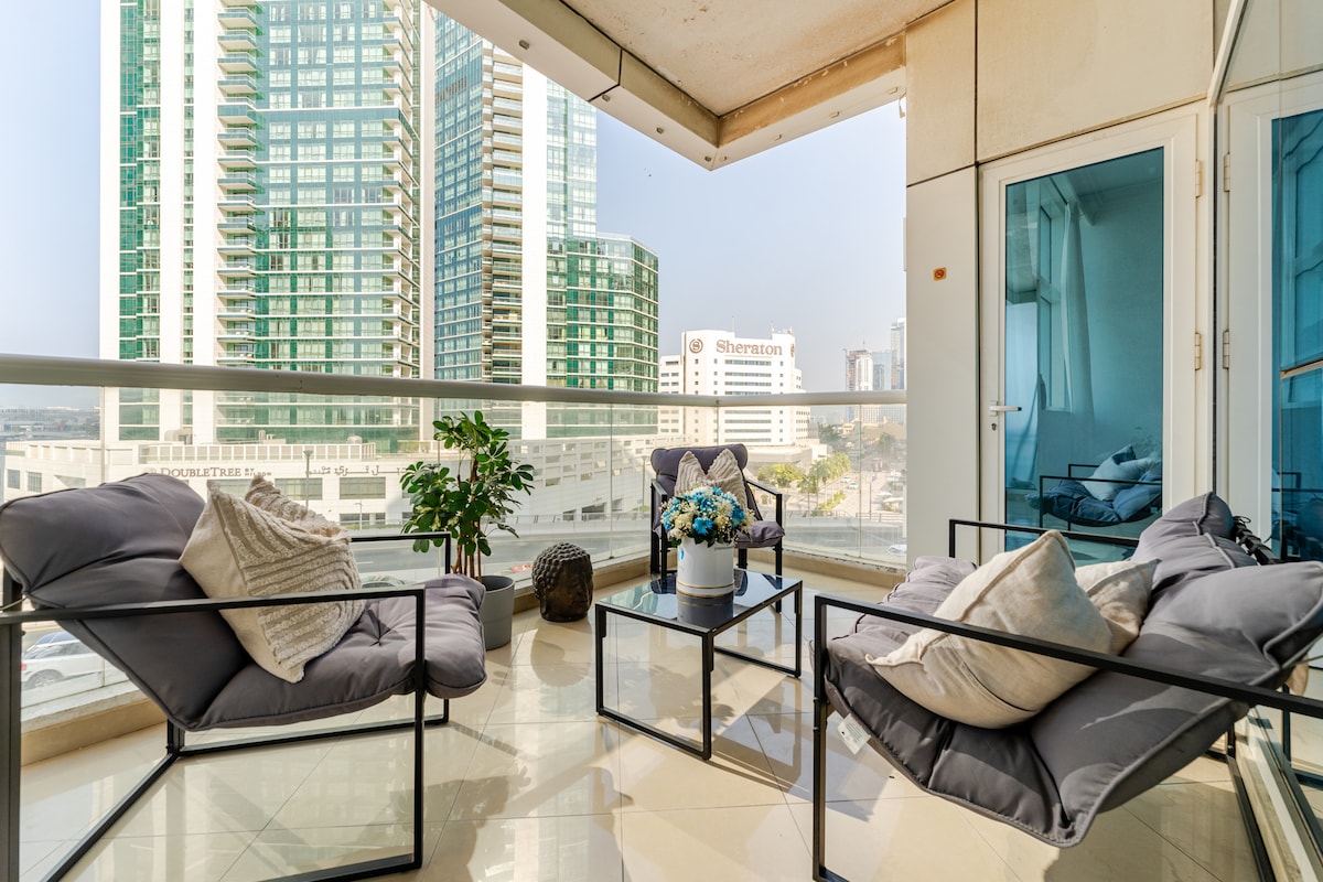A modern balcony is furnished with comfortable seating arrangements consisting of plush armchairs and a small table. Potted plants add a touch of greenery, while high-rise buildings are visible in the background, reflecting the urban setting.