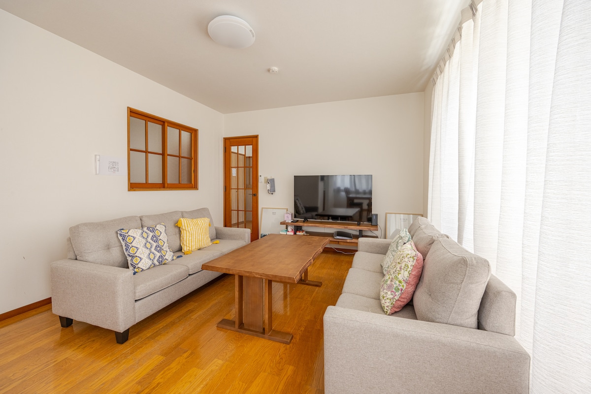 A comfortable living area is furnished with two gray sofas and a wooden coffee table. A large television is positioned on a console, while natural light enters through sheer curtains framing the window. A cozy ambience is created by decorative pillows on the sofas.