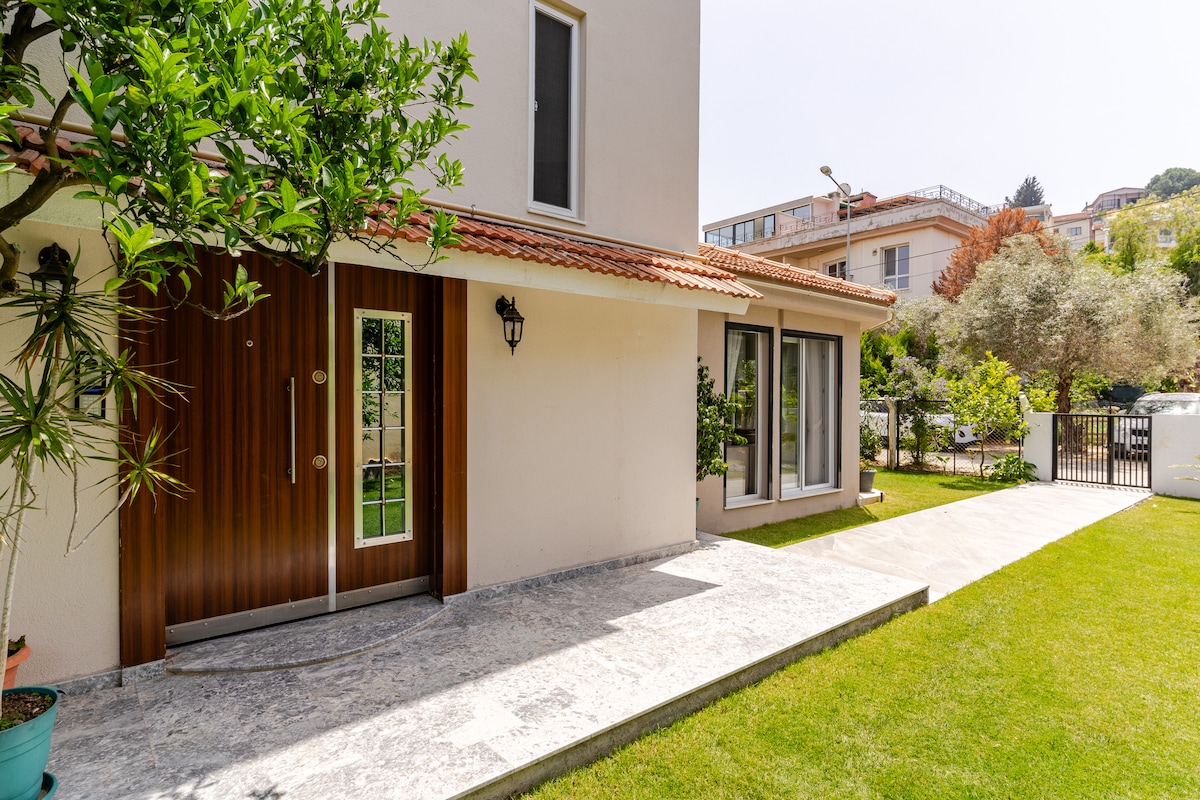 The exterior of the house features a landscaped garden with lush green grass. A modern entryway is highlighted by double doors with glass panels. A smooth stone pathway leads to the entrance, complemented by surrounding plants and trees that provide a serene atmosphere.