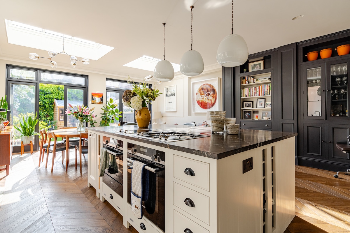 An open-plan kitchen is revealed, featuring a large island with a dark countertop. Natural light floods the space through skylights and windows. Modern pendant lights are suspended above the island, while plants provide a fresh touch. Dining and seating areas are seen in the background.