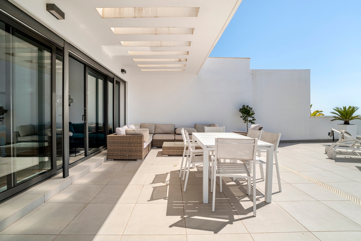 A spacious terrace is furnished with a sectional sofa and a dining area. Large glass doors allow natural light to flow in, while potted plants add greenery. The area is designed for relaxation and outdoor dining, with an unobstructed view of the clear blue sky.