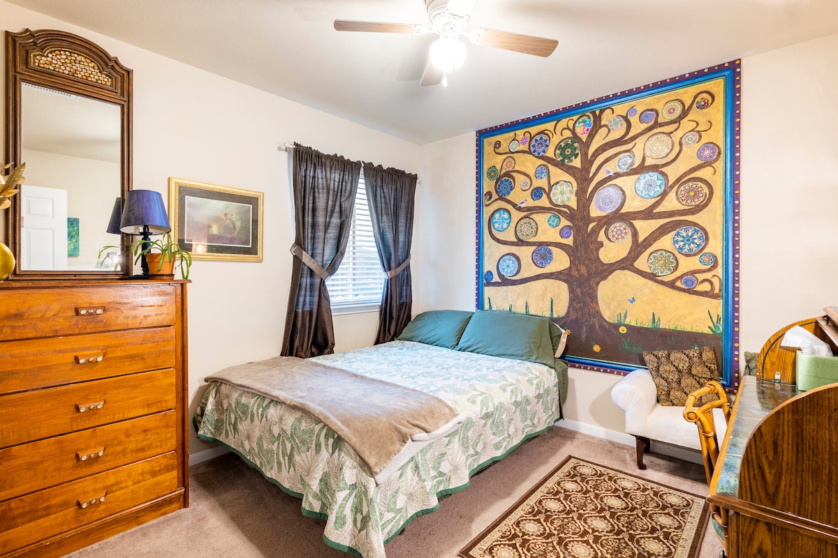 A cozy bedroom features a spacious bed with green and white bedding. A striking wall mural of a colorful tree adorns one side, while a mirror, dresser, and writing desk provide functional furnishings. Natural light filters through two elegantly draped windows.