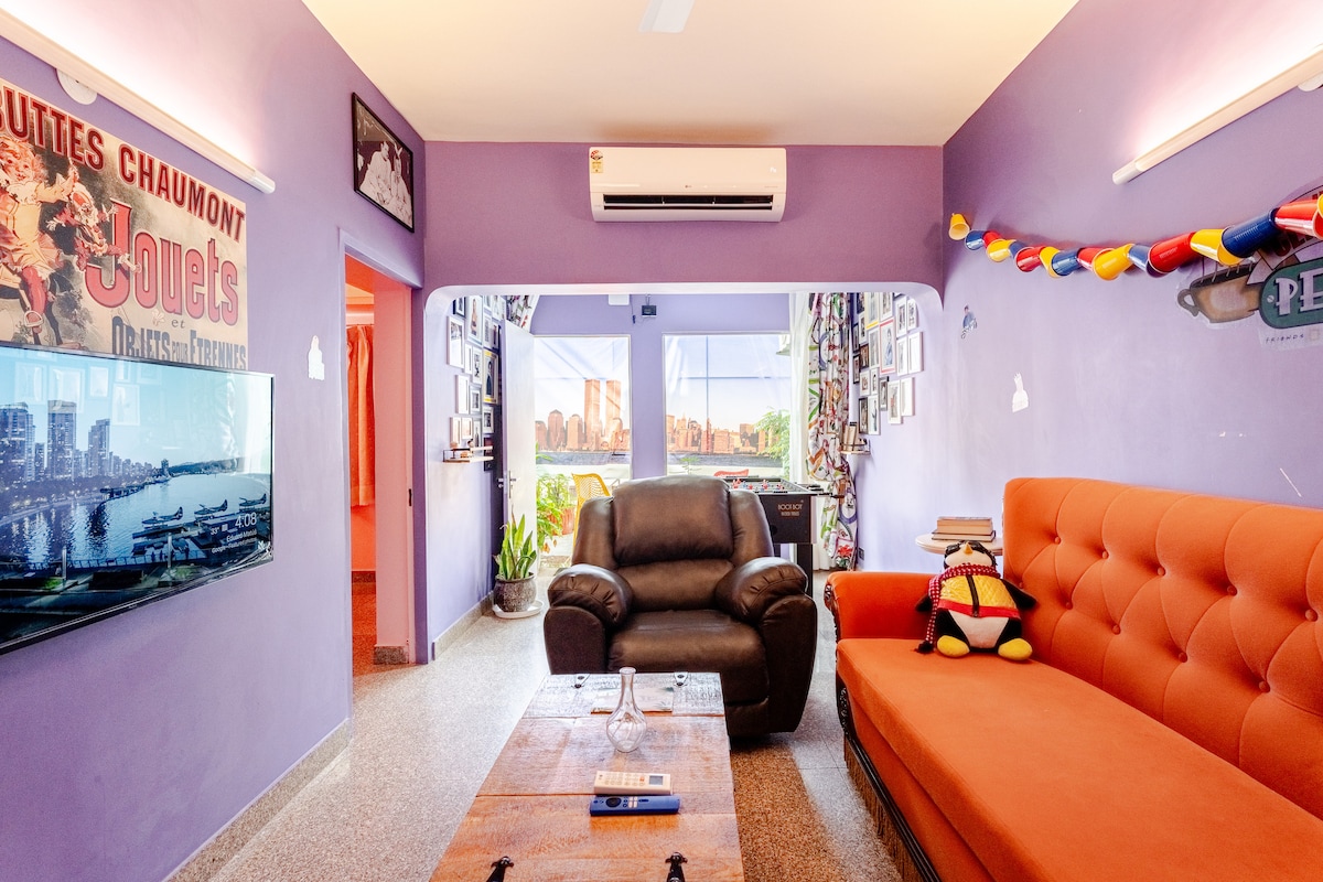 The living area is characterized by a vibrant purple accent wall. An orange couch and dark recliner chair are positioned around a wooden coffee table. A wall-mounted TV is visible, and colorful decorative elements add energy to the space.