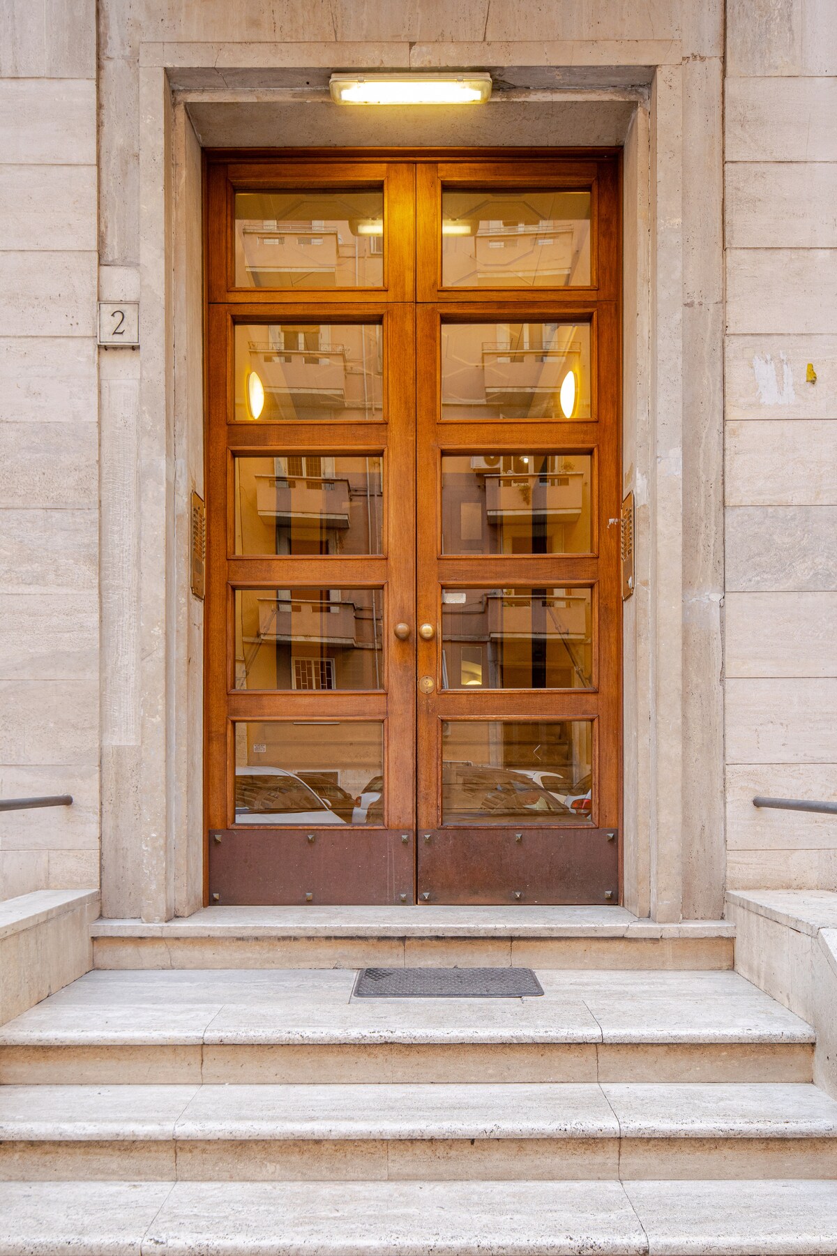 The entrance features a wooden double door with large glass panels, providing a view into the hallway beyond. Stone steps lead up to the doors, framed by a light-colored stone facade. Soft illumination highlights the entryway, creating an inviting atmosphere.