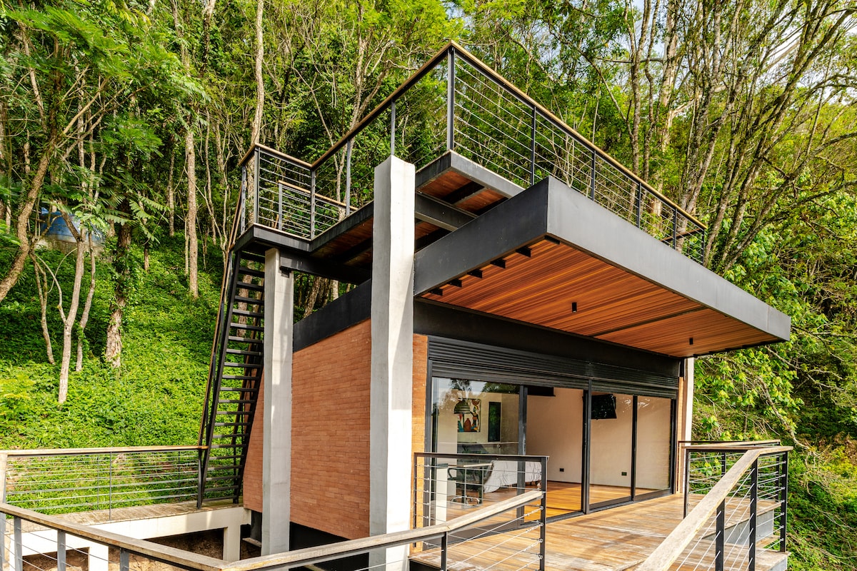 A modern two-story chalet is surrounded by lush greenery, featuring an expansive balcony on the upper level. The exterior showcases a blend of brick and stone elements, while large glass doors provide a connection to the natural surroundings.