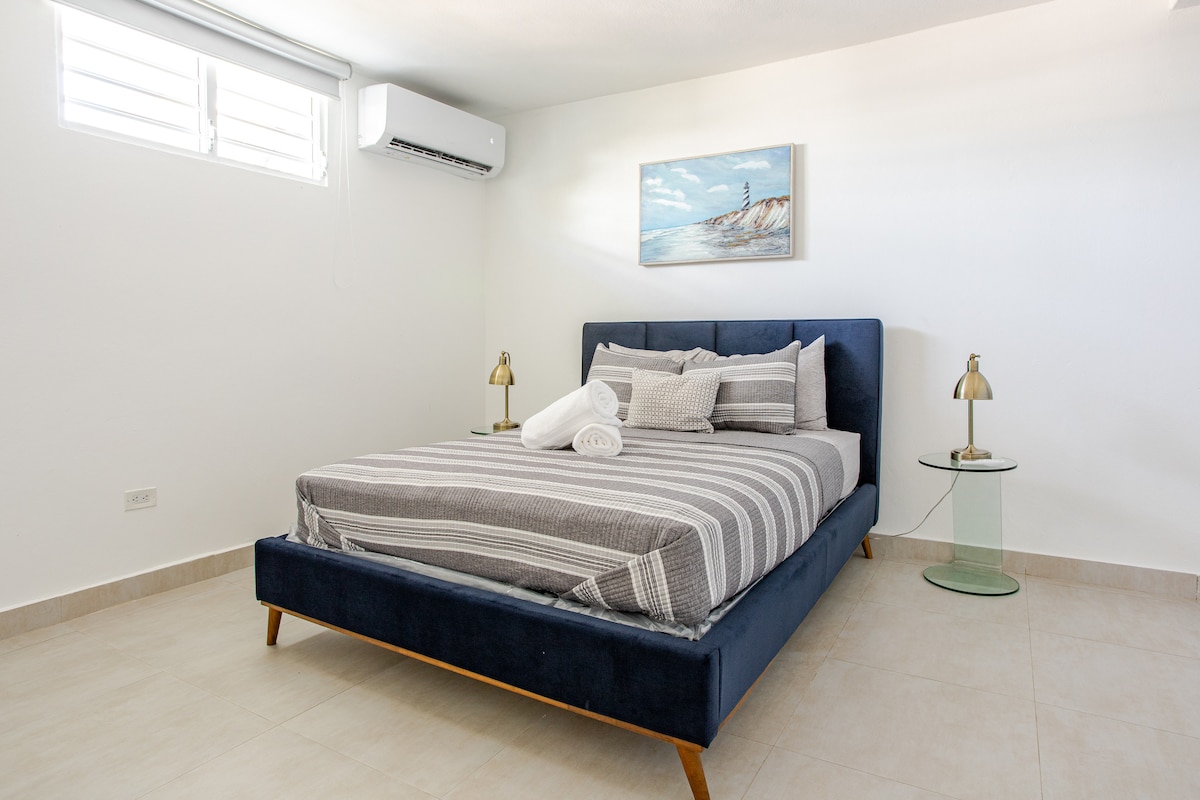 A spacious bedroom features a navy blue bed frame with a striped comforter and decorative pillows. Two glass side tables with lamps sit on either side of the bed. A piece of art depicting a beach scene adorns the wall. Natural light enters through a window with shutters.