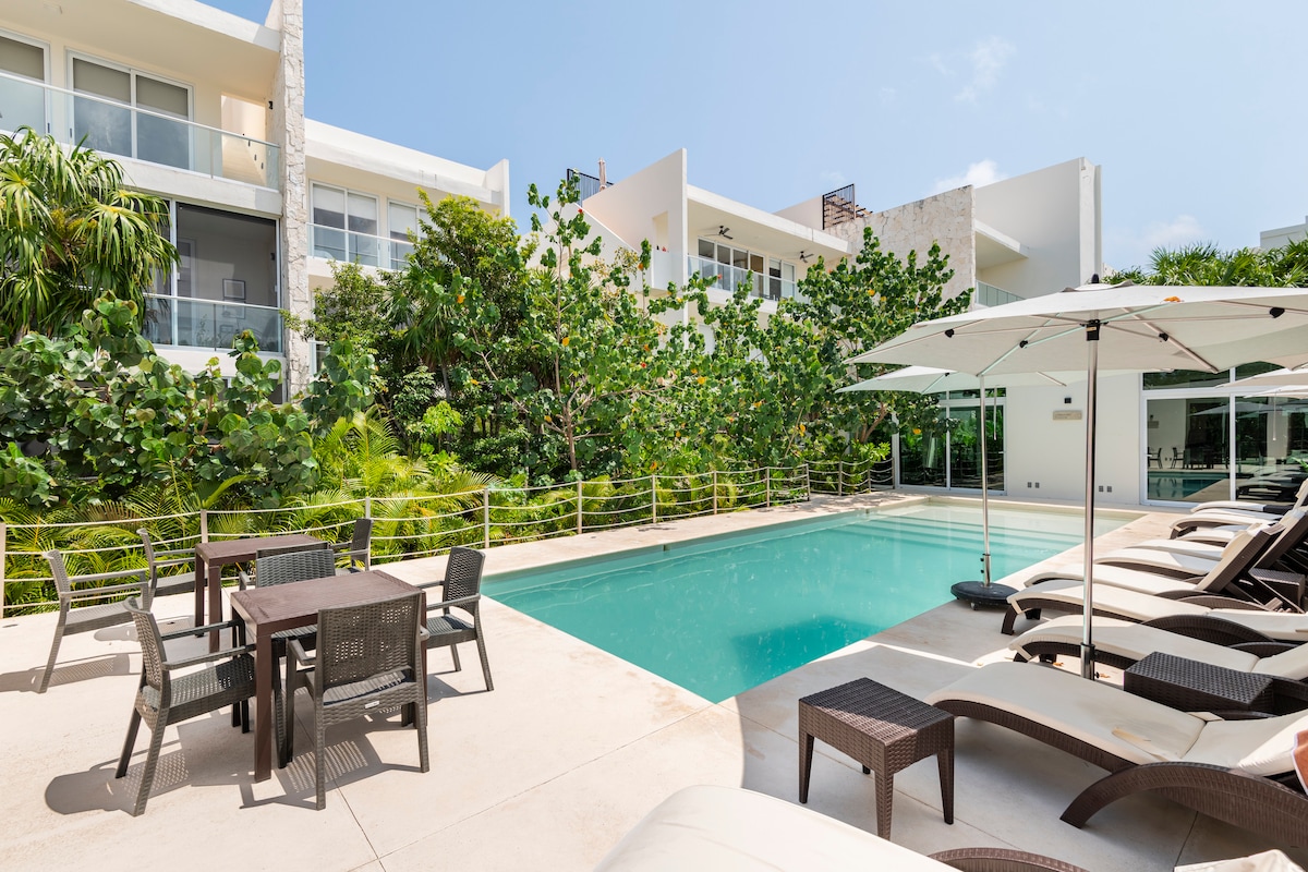 A modern pool area is surrounded by lush greenery, featuring a clear blue pool with lounge chairs and an umbrella nearby. A dining table with chairs is positioned adjacent to the water, complementing the serene atmosphere of the outdoor space.