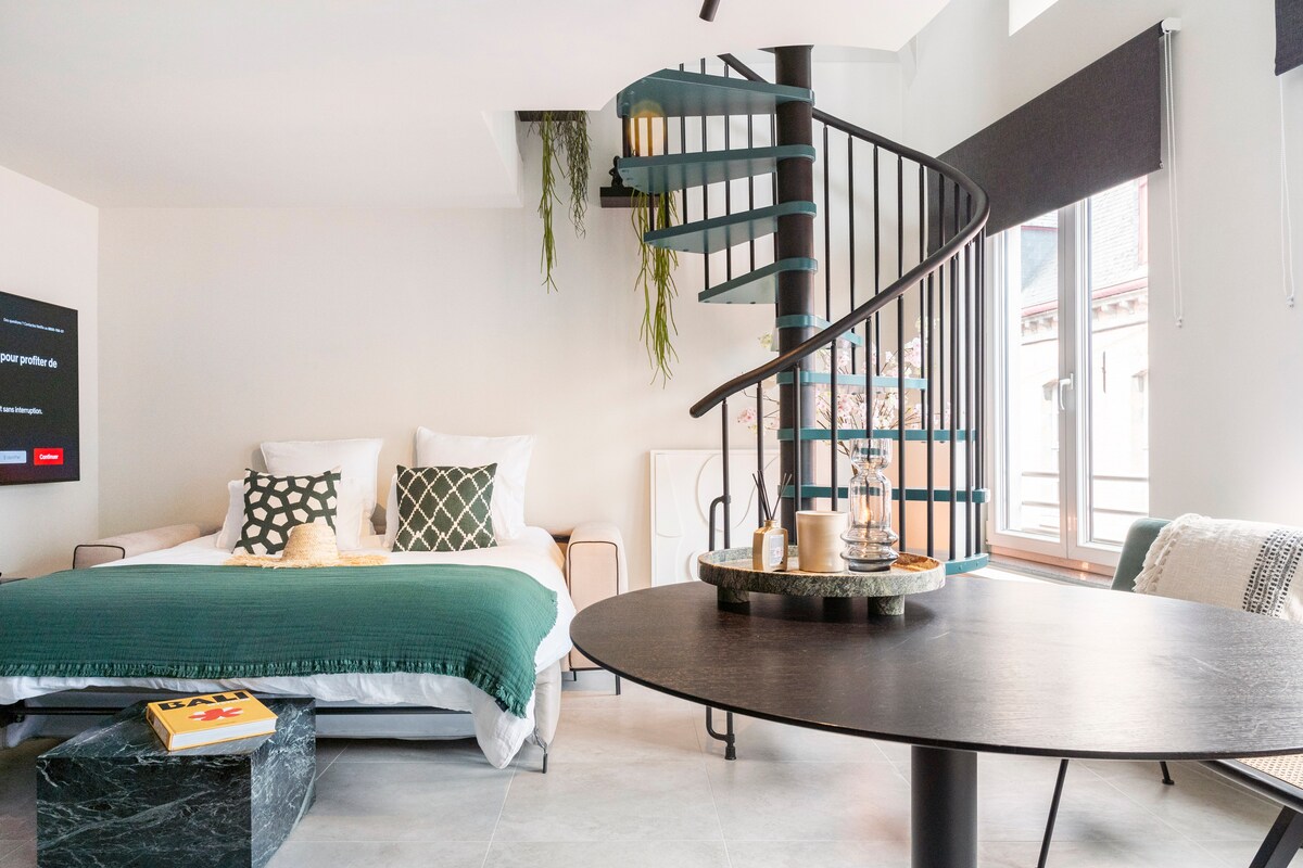 A modern living space features a spiral staircase leading to an upper level. A spacious bed is arranged with decorative pillows and a textured blanket. A round dining table with a dark top is positioned in the foreground, complemented by a minimalist decor.