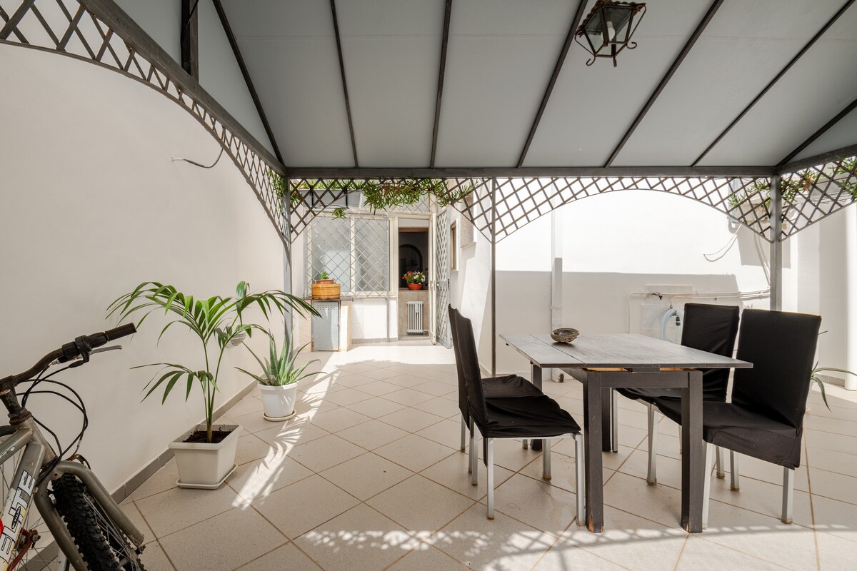 An outdoor gazebo area is presented with a table and black chairs, suitable for meals or relaxation. A potted plant is placed nearby, adding a touch of greenery. The entrance to the interior is visible in the background, framed by light-colored walls.