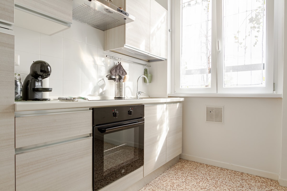 A modern kitchen is equipped with a sleek countertop and cabinetry in light wood tones. A black oven is visible below a built-in stovetop, while a coffee machine is placed nearby. Natural light filters through a large window, illuminating the space.