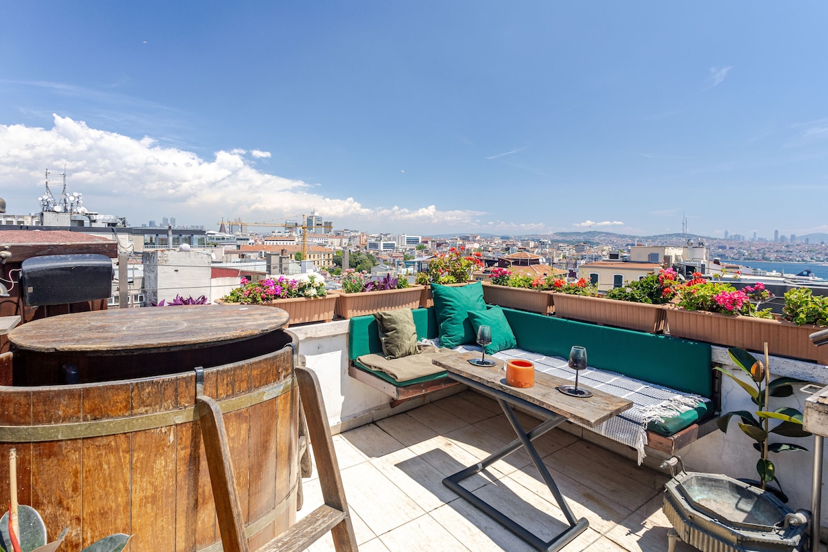A spacious rooftop terrace features a wooden hot tub and a seating area surrounded by lush flowers. The view encompasses the city skyline and the Bosphorus, with comfortable cushions placed along the edge for relaxation. The bright blue sky creates an inviting atmosphere.