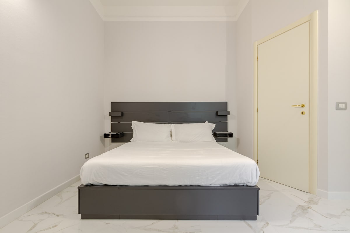 A spacious bedroom features a contemporary bed with minimalistic side tables. The light-colored walls and elegant marble floor contribute to a serene environment. A door is visible, leading to the rest of the apartment, ensuring an inviting space for relaxation.