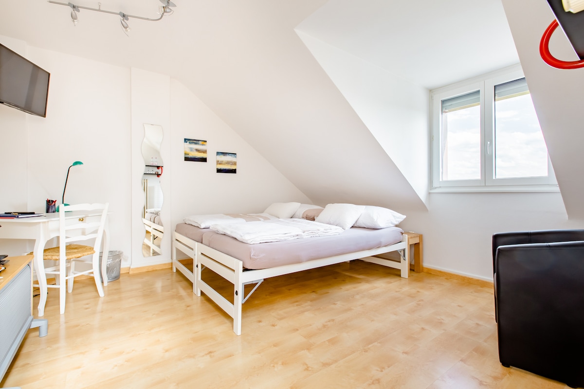 A bright attic room features a spacious bed with crisp white linens and two pillows. A small desk with a green lamp is positioned beside a mirror. Natural light enters through a window, offering a view of the sky. Light wood flooring adds warmth.