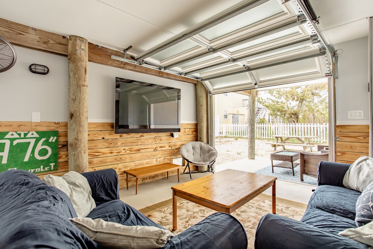 An indoor recreational space features a wall-mounted television and a comfortable seating area with an inviting coffee table. Large garage doors provide access to a patio area with outdoor seating, allowing for a seamless transition between indoor and outdoor relaxation.