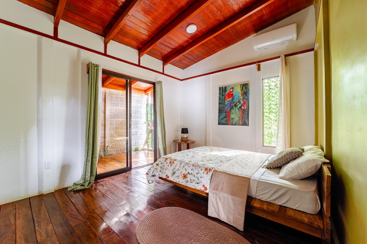 A spacious bedroom features hardwood flooring and a king-sized bed topped with neutral linens. Sunlight filters through large glass doors, which lead to a private balcony. A vivid painting of a tropical bird adorns one wall, complemented by soft green curtains and a bedside lamp.