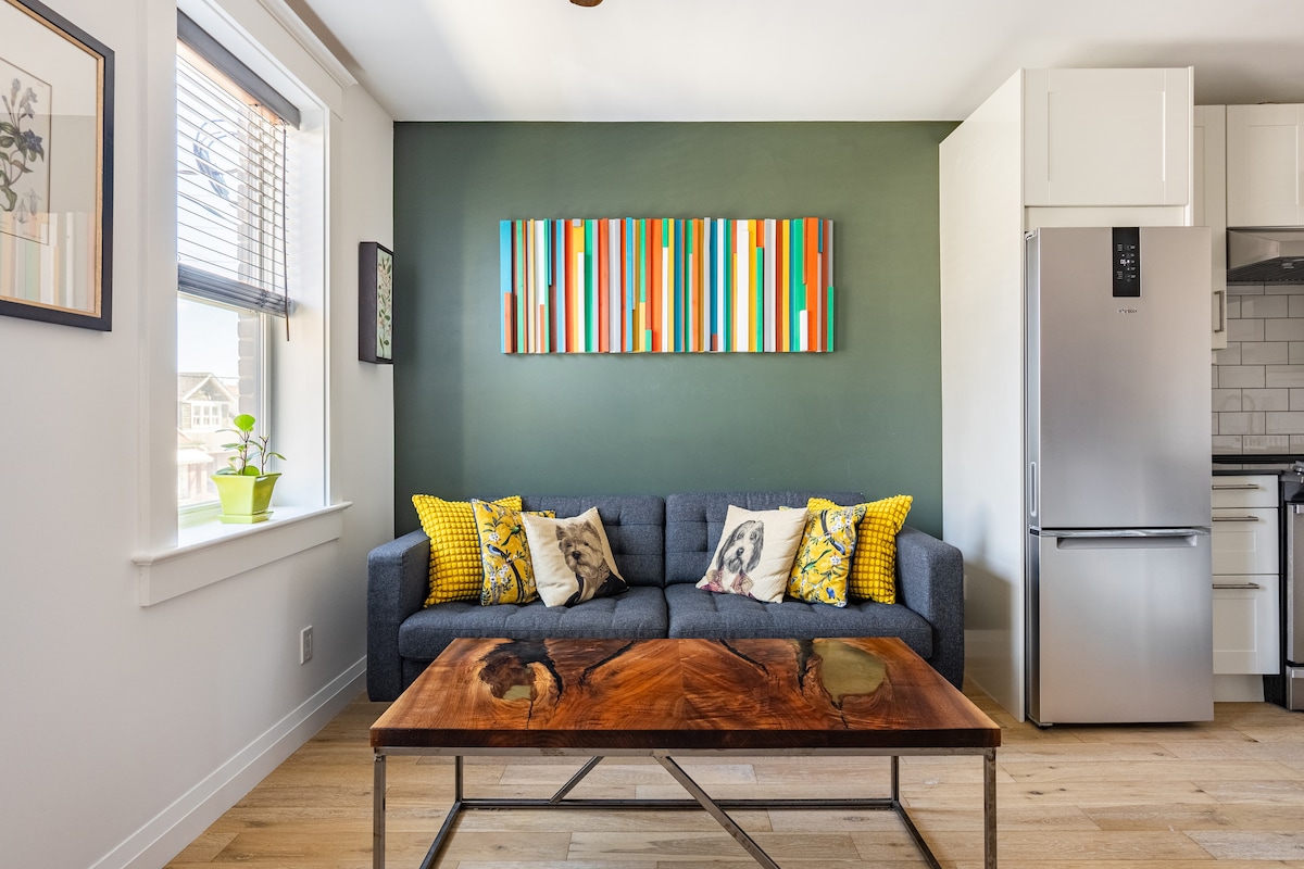 A modern living space features a grey sofa adorned with patterned throw pillows. A rustic wooden coffee table sits in front, complemented by a vibrant wall art piece above. A stainless steel refrigerator and kitchen area are visible in the background, with ample natural light from the windows.