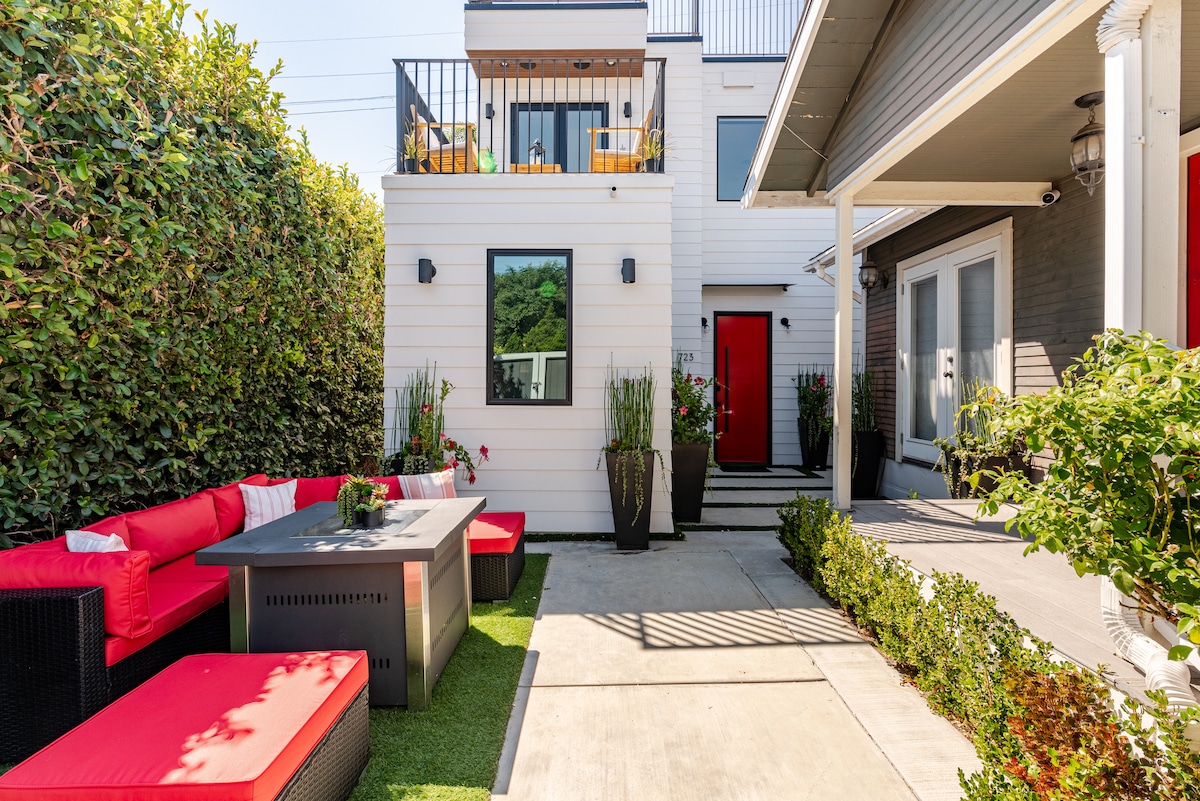 An outdoor patio area is showcased, featuring a modern stone table surrounded by red seating. Lush greenery lines the space, enhancing the privacy. A bright red door adds a pop of color to the exterior, with sunlight spilling across the concrete pathway.