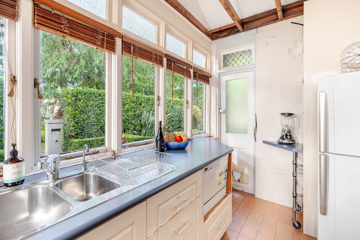 A functional kitchen is illuminated by natural light from large windows overlooking a garden. The space includes a double sink, a stove below counter, and a refrigerator. A small table in the corner provides additional surface area, complementing the overall layout.