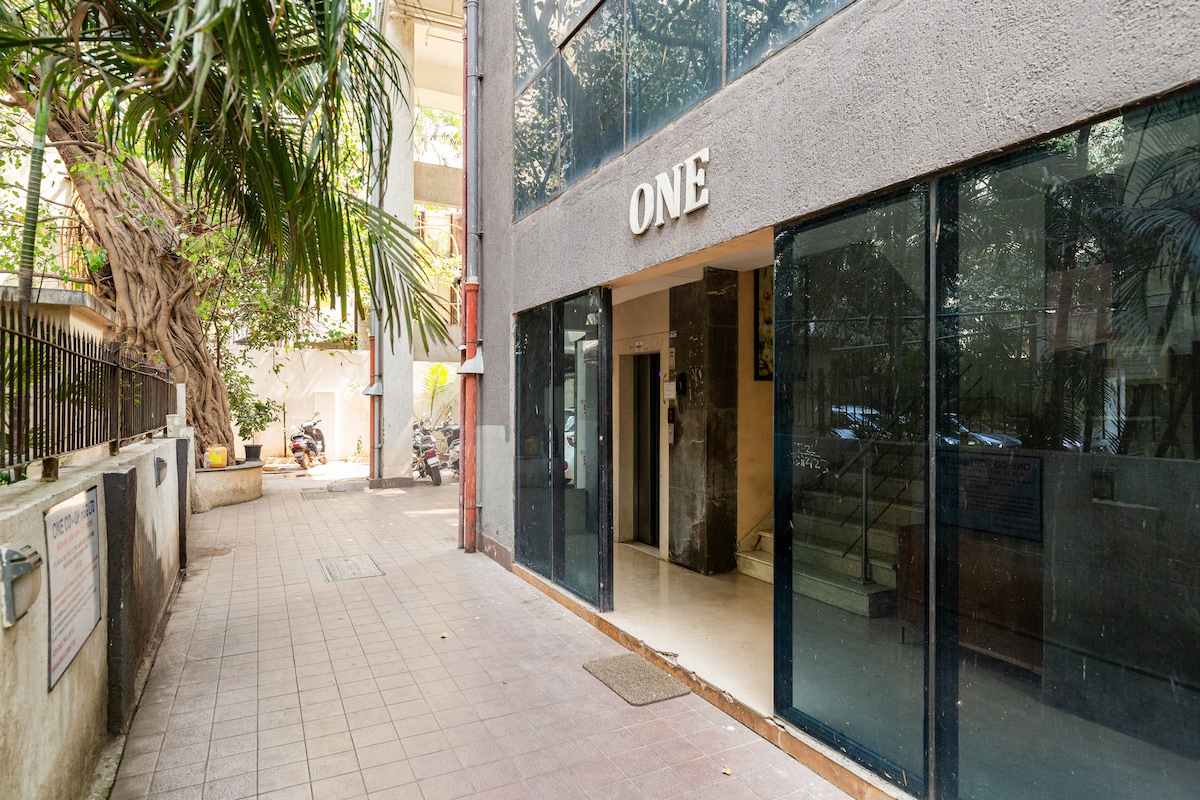 The entrance of the modern building is showcased, featuring large glass doors that provide a clear view of the interior lobby. A tiled pathway leads to the entrance, framed by lush greenery on either side, enhancing the welcoming environment.