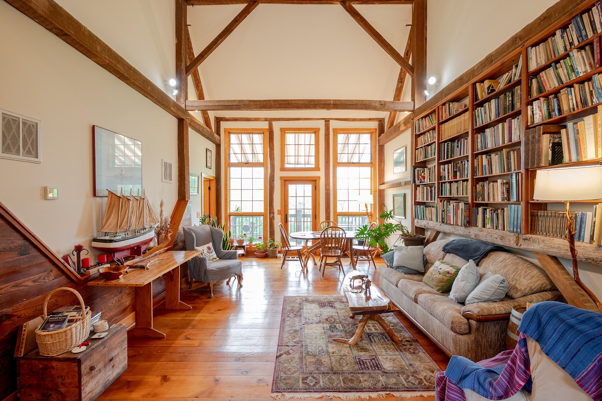 A spacious living area showcases high wooden beams and large windows allowing natural light. A cozy sofa is arranged with decorative cushions, while books line the walls. A dining table is positioned to one side, with a variety of plants adding a touch of greenery.