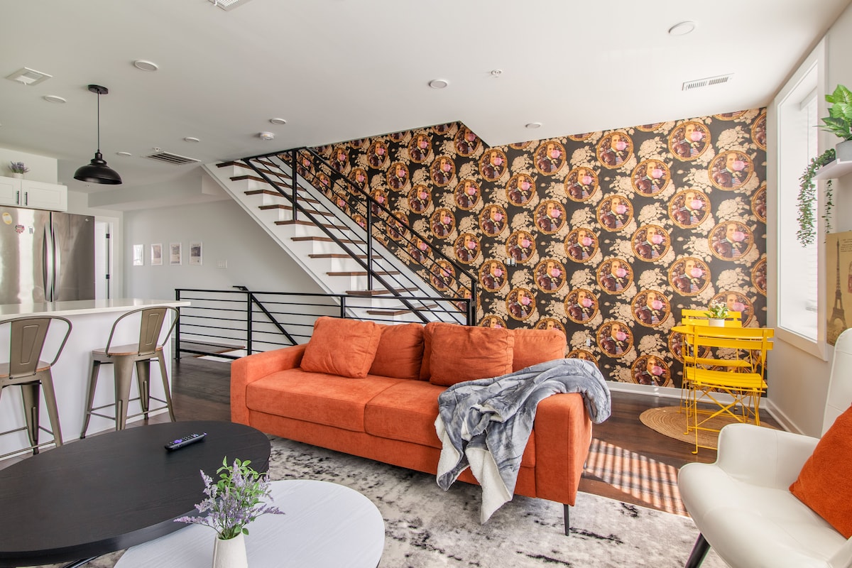 A bright living area features an orange sectional sofa adorned with a gray throw. A black coffee table sits in front, while a vibrant patterned wall adds character. The open layout allows views of the modern kitchen and a stylish staircase leading to the upper level.