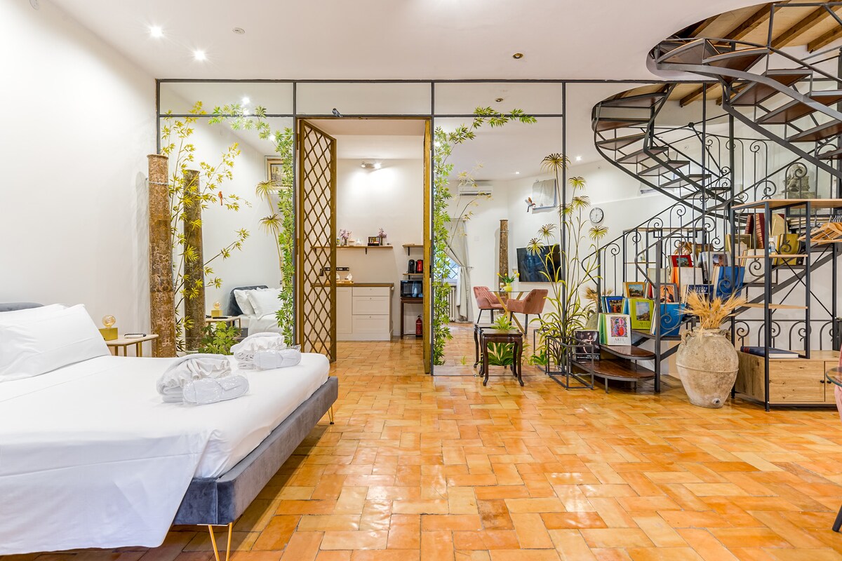 An open living space features a spacious bed with fresh linens and towels. Glass walls border the room, allowing natural light to illuminate the terracotta flooring. A spiral staircase leads to the upper level, and greenery adds a touch of nature throughout the area.
