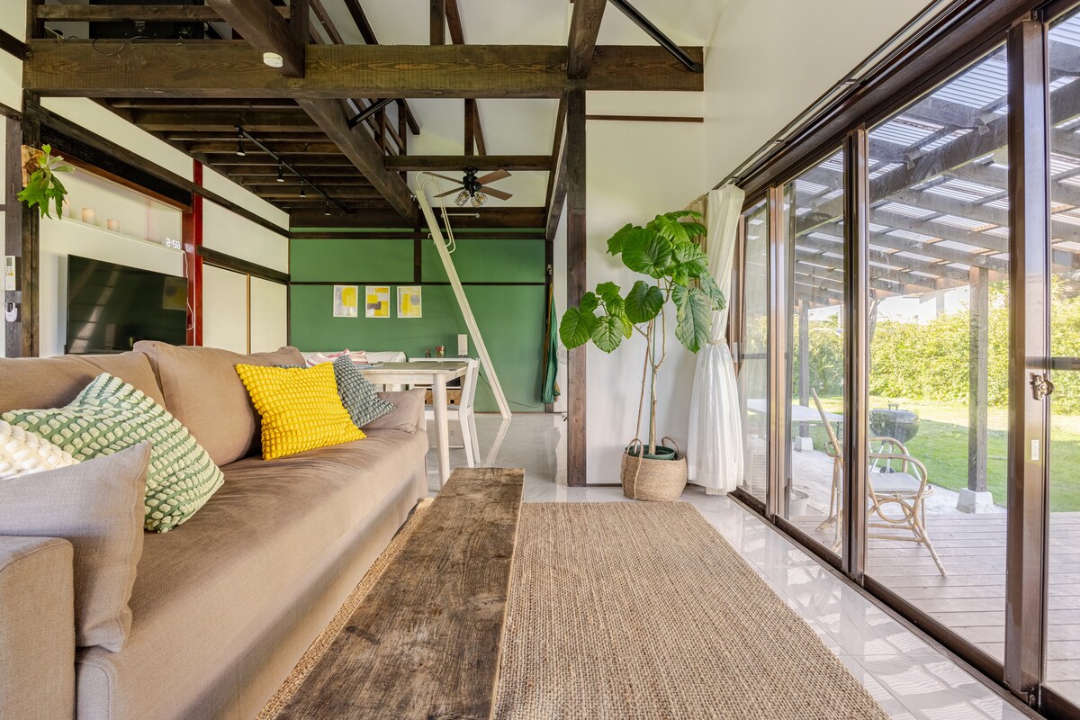 The spacious living area features natural wood beams and large glass doors that open to a green garden. A comfortable sofa is paired with decorative cushions, and a neutral rug is laid on the tiled floor. A dining table is visible in the background.