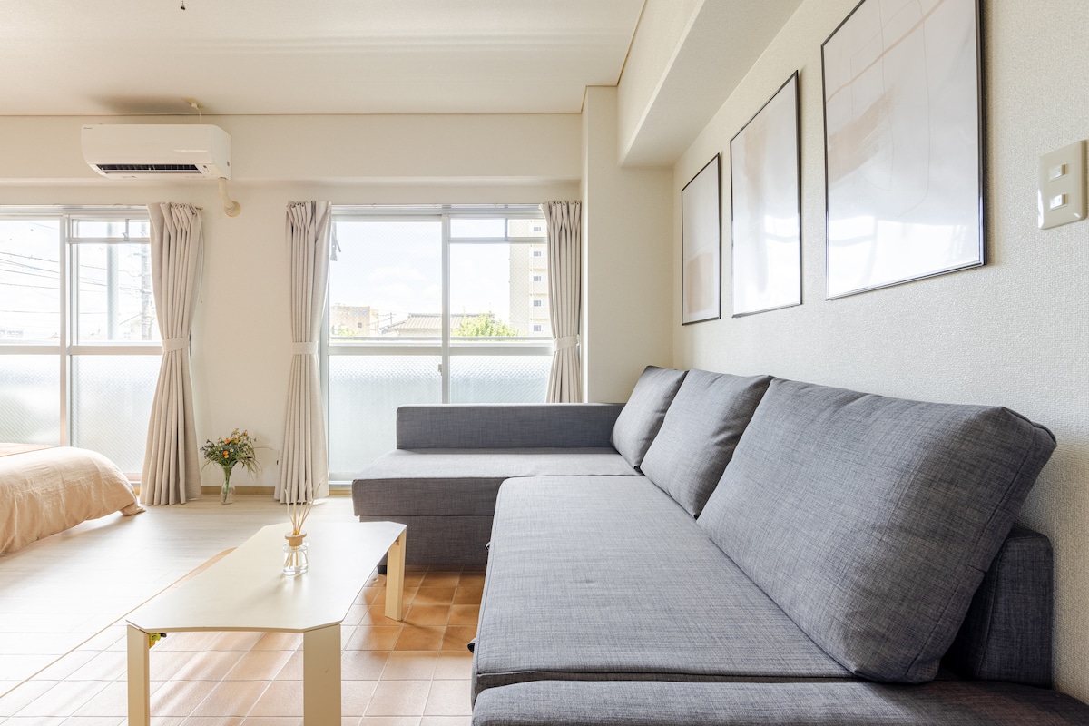 A comfortable seating area is arranged with a spacious grey sofa and a low coffee table. Natural light filters through large windows with soft curtains. Framed artwork is displayed on the wall above the sofa, contributing to a pleasant environment.