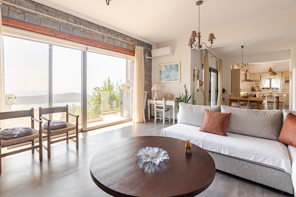 A spacious salon featuring large glass doors that open to scenic views. A round wooden table sits at the center, accompanied by a modern sofa with cushions. Two wooden chairs are arranged near the window, enhancing the room's inviting setup. The kitchen is visible in the background.