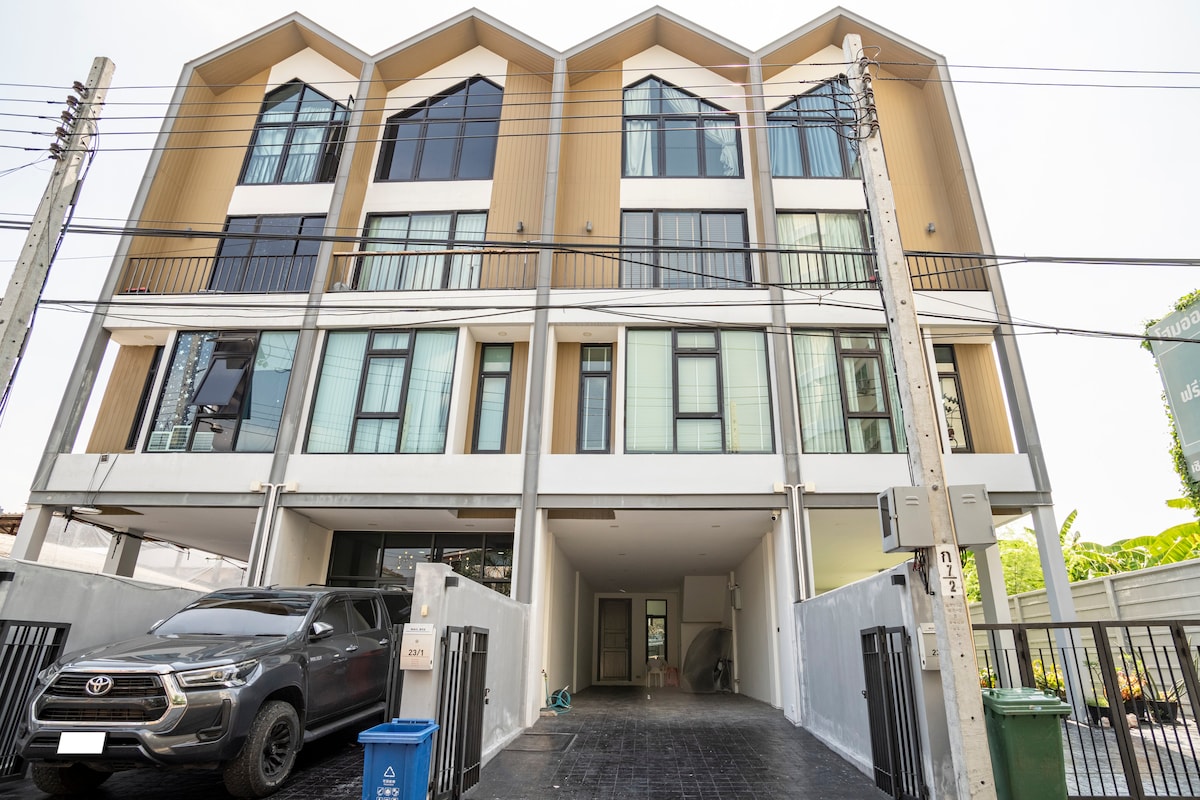 The property features a modern, multi-story building with a distinctive architectural design. Large windows allow ample natural light, while a spacious driveway accommodates vehicles. A blue recycling bin and an outdoor light fixture are visible at the front entrance.