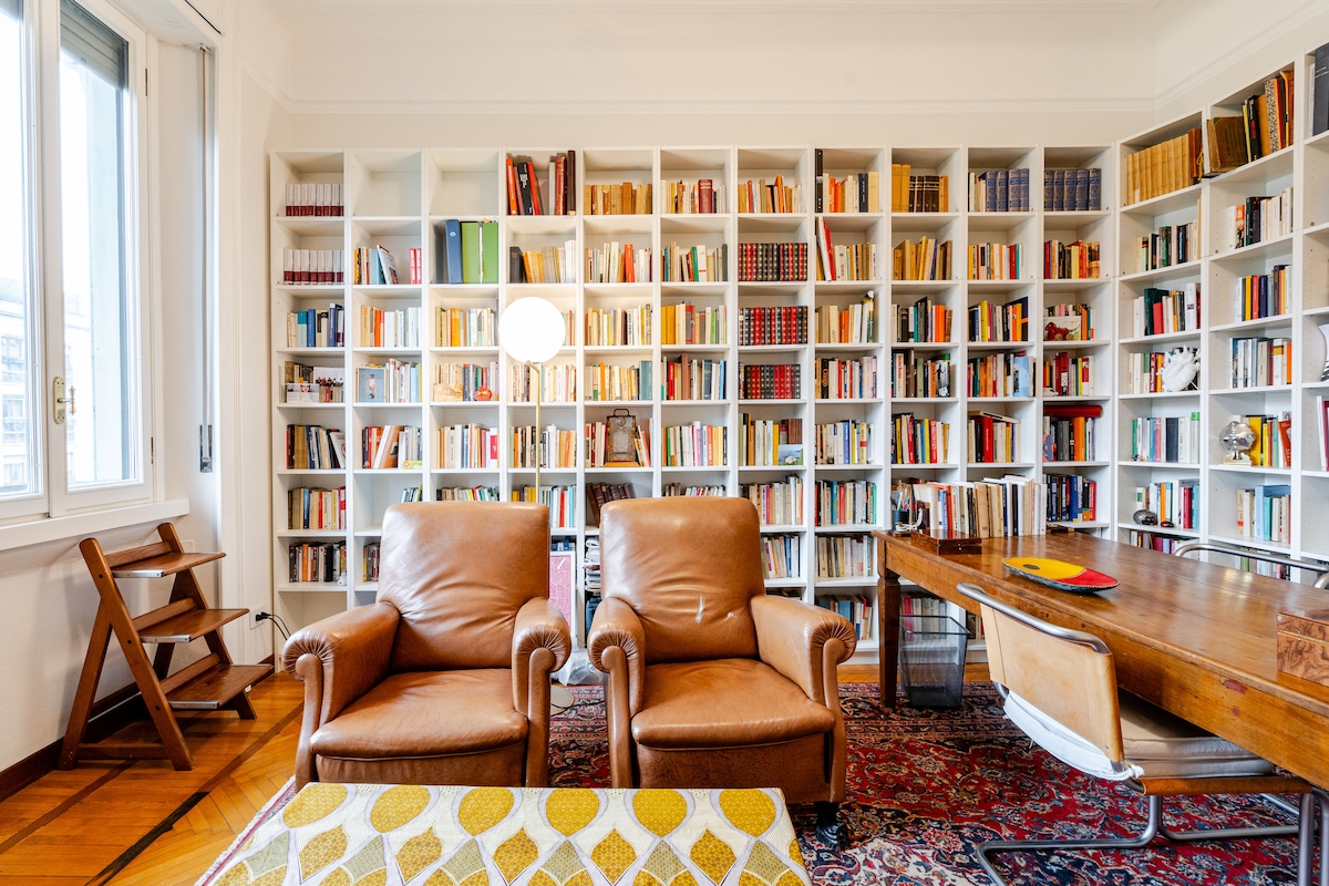 A bright study area features two leather armchairs positioned near a wooden table. A wall of bookshelves fills the space, displaying an extensive collection of books in various colors. Natural light enters through the window, enhancing the warm tones of the room.