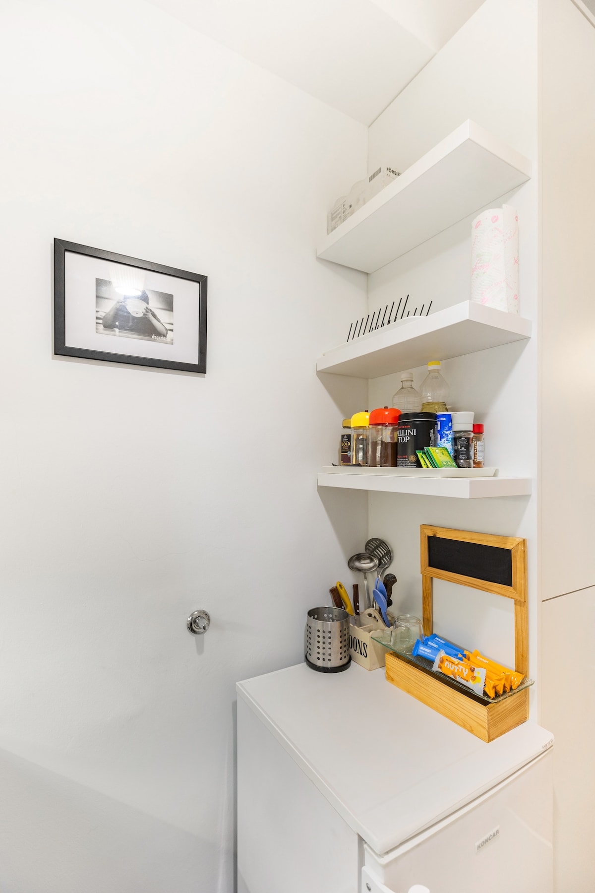 A compact kitchen area is showcased, featuring open shelves holding various kitchen supplies and condiments. A small refrigerator is placed beneath the shelves, while a wooden basket containing utensils and packaged goods sits on a counter. A framed photograph adds a decorative touch to the wall.