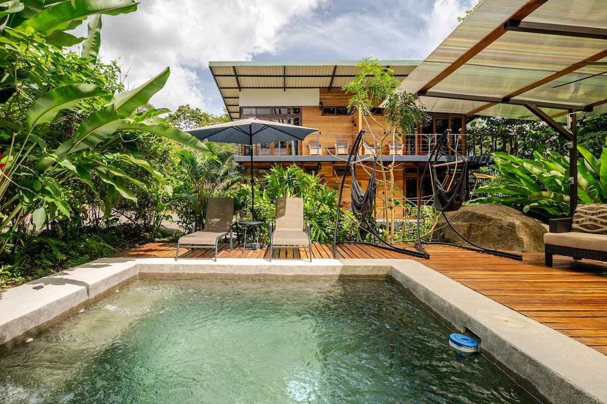A serene outdoor space features a refreshing pool with clear water, surrounded by lush greenery. Comfortable loungers, shaded by an umbrella, are positioned nearby, while swinging chairs add a unique touch. The two-story wooden structure looms in the background, surrounded by tropical foliage.