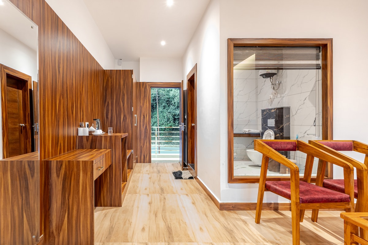 The interior space features a wooden decor with vibrant accents. A seating area is highlighted by two wooden chairs with plush cushions. A kitchenette with a kettle is positioned nearby, while a window offers a view of the outdoors. A glass panel reveals a modern bathroom.