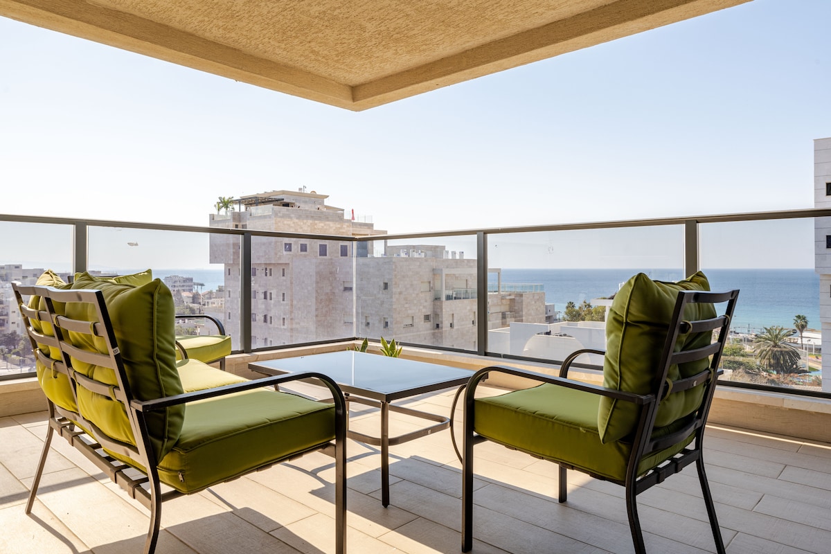 A terrace area is depicted featuring two green lounge chairs flanking a glass-top table. In the background, the ocean can be seen beyond low-rise buildings, with clear skies overhead, suggesting a relaxing outdoor space.