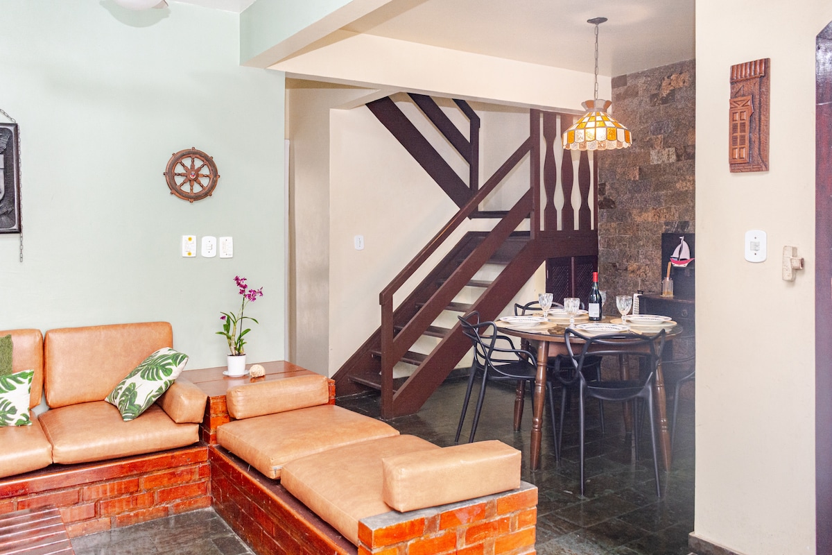 An integrated living and dining area is presented, featuring a comfortable brown leather seating arrangement. A wooden dining table with chairs is visible, alongside a decorative wall with natural stone accents. A staircase leads to the upper level, enhancing the home's spaciousness.