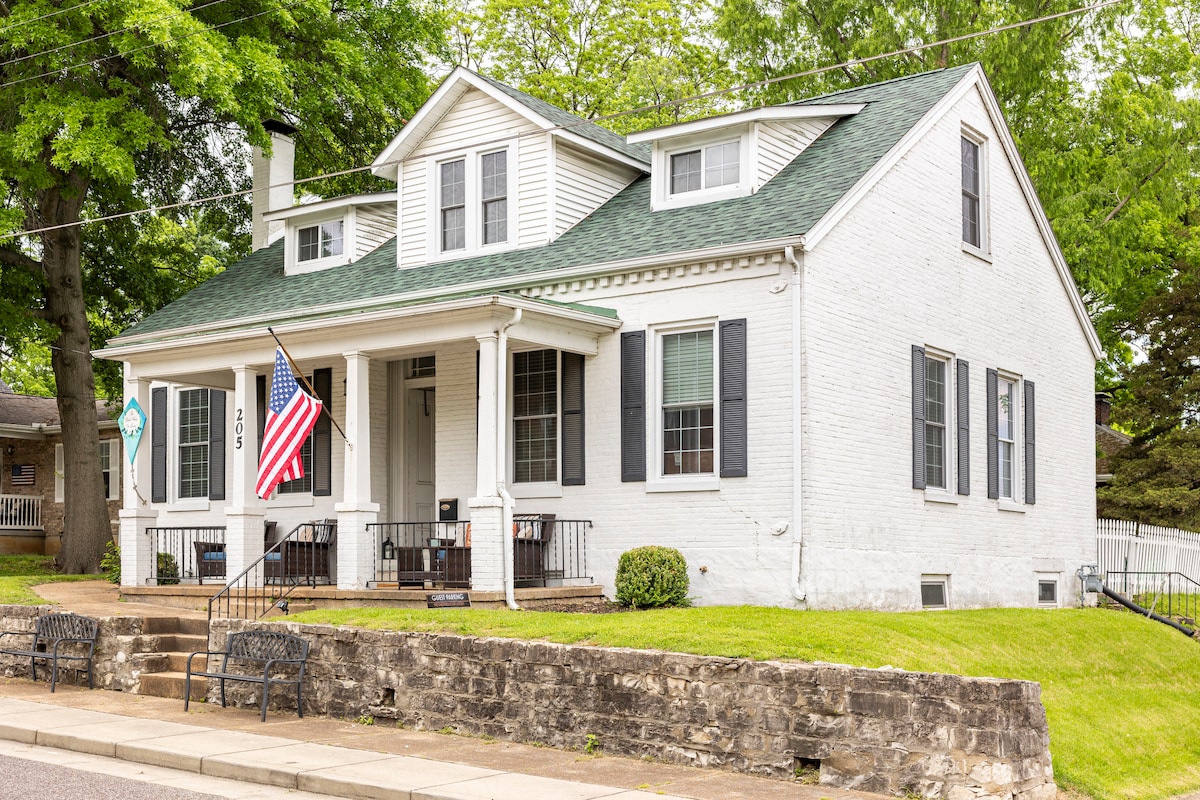 The charming exterior of a large white house is shown, featuring a gabled roof and a welcoming front porch. American flags are displayed, while well-maintained green lawns and stone steps lead to the entrance. Trees provide natural shade around the property.