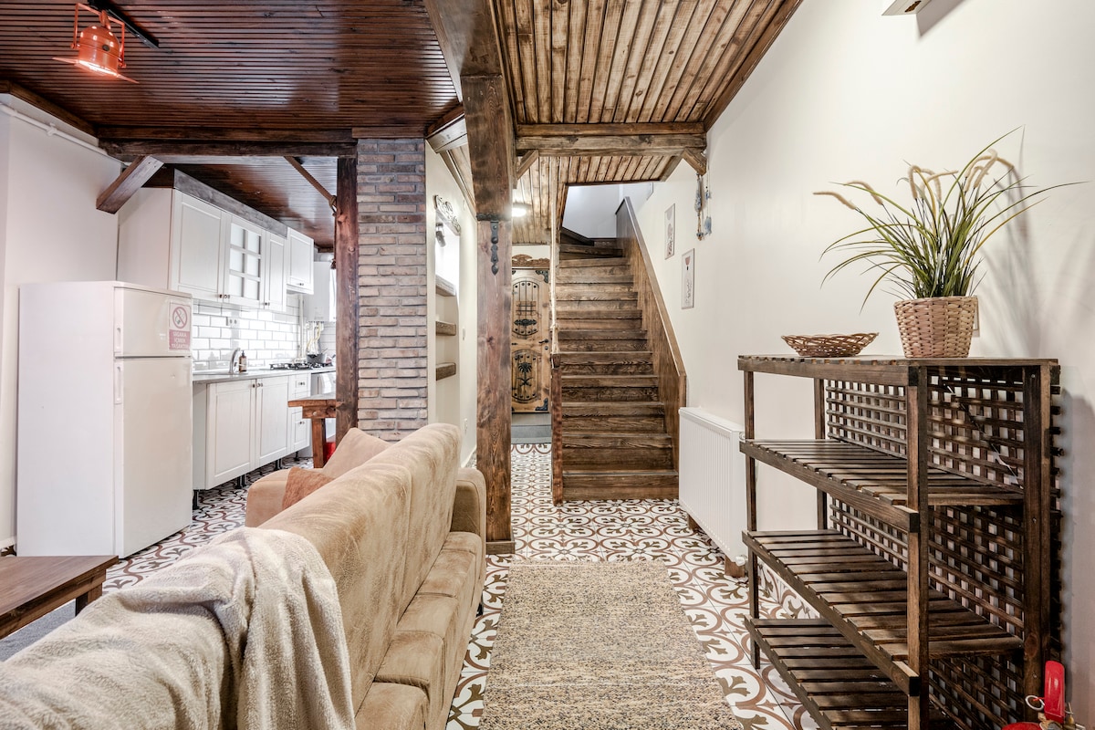 An open living area features a comfortable sofa and a textured rug on colorful tiled flooring. A wooden staircase leads to the upper level, while a modern kitchen area is visible in the background, equipped with essential appliances. Natural light illuminates the space.
