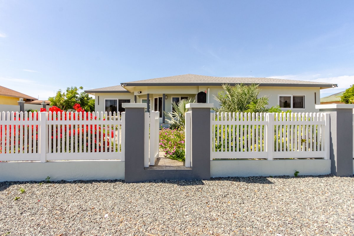 A single-story house is framed by a white picket fence, with colorful flowering plants lining the walkway. The façade features large windows that bring in natural light, and a spacious front porch is visible, creating an inviting entrance.