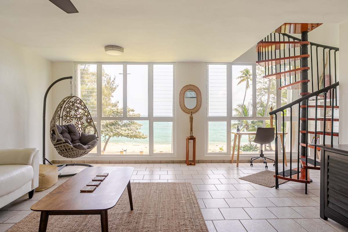 A well-lit living area features large windows showcasing a view of the ocean and palm trees. A cozy hanging chair is positioned near the window, while a round mirror and a small desk with a chair are also present. A spiral staircase leads to the upper level.