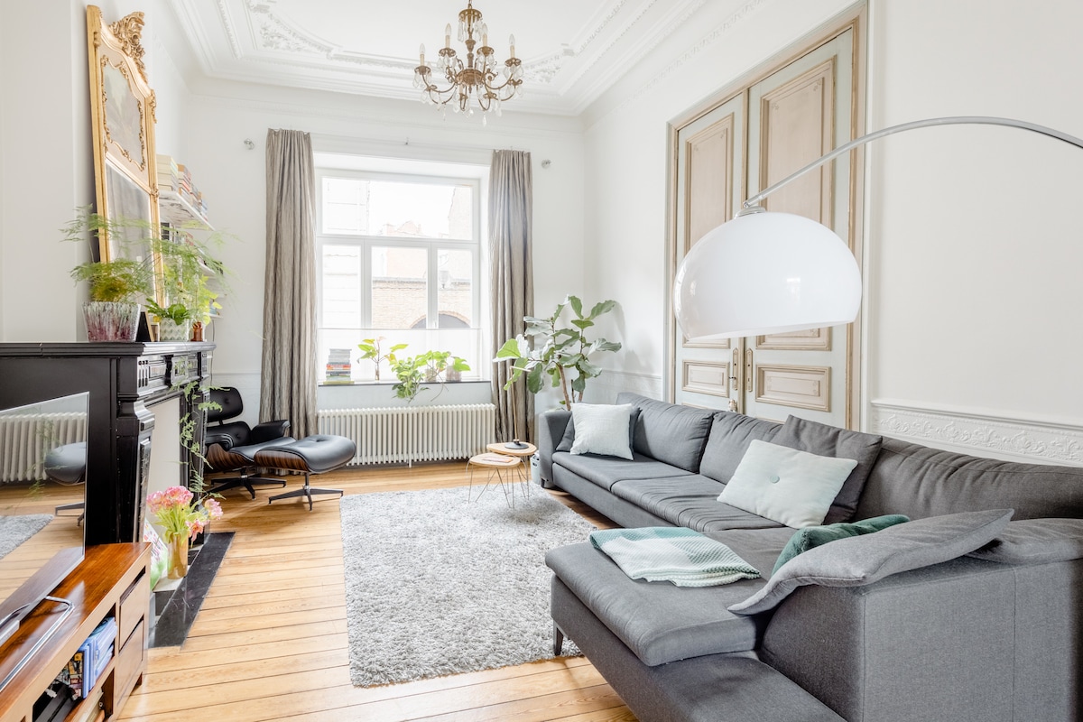 A spacious living area features a large grey sectional sofa and a light-wood coffee table. A round rug covers the hardwood floor, while potted plants add greenery. Tall windows with light curtains illuminate the room, and a decorative chandelier hangs from the ceiling.