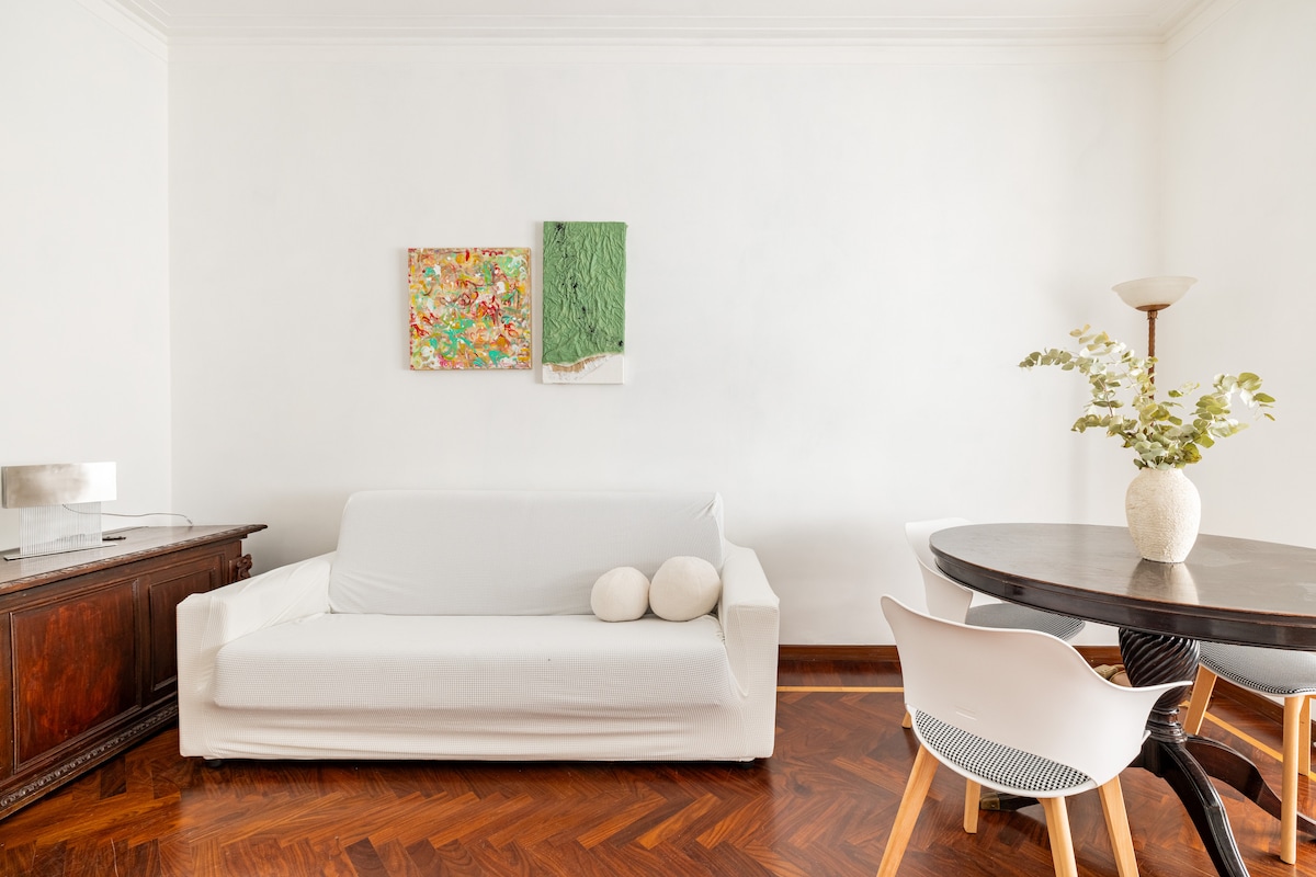 A bright and inviting living space features a white sofa adorned with two cushions. A small round dining table, paired with modern chairs, sits nearby. Colorful artwork is displayed on the wall, and a lamp stands beside a vintage cabinet on the left.