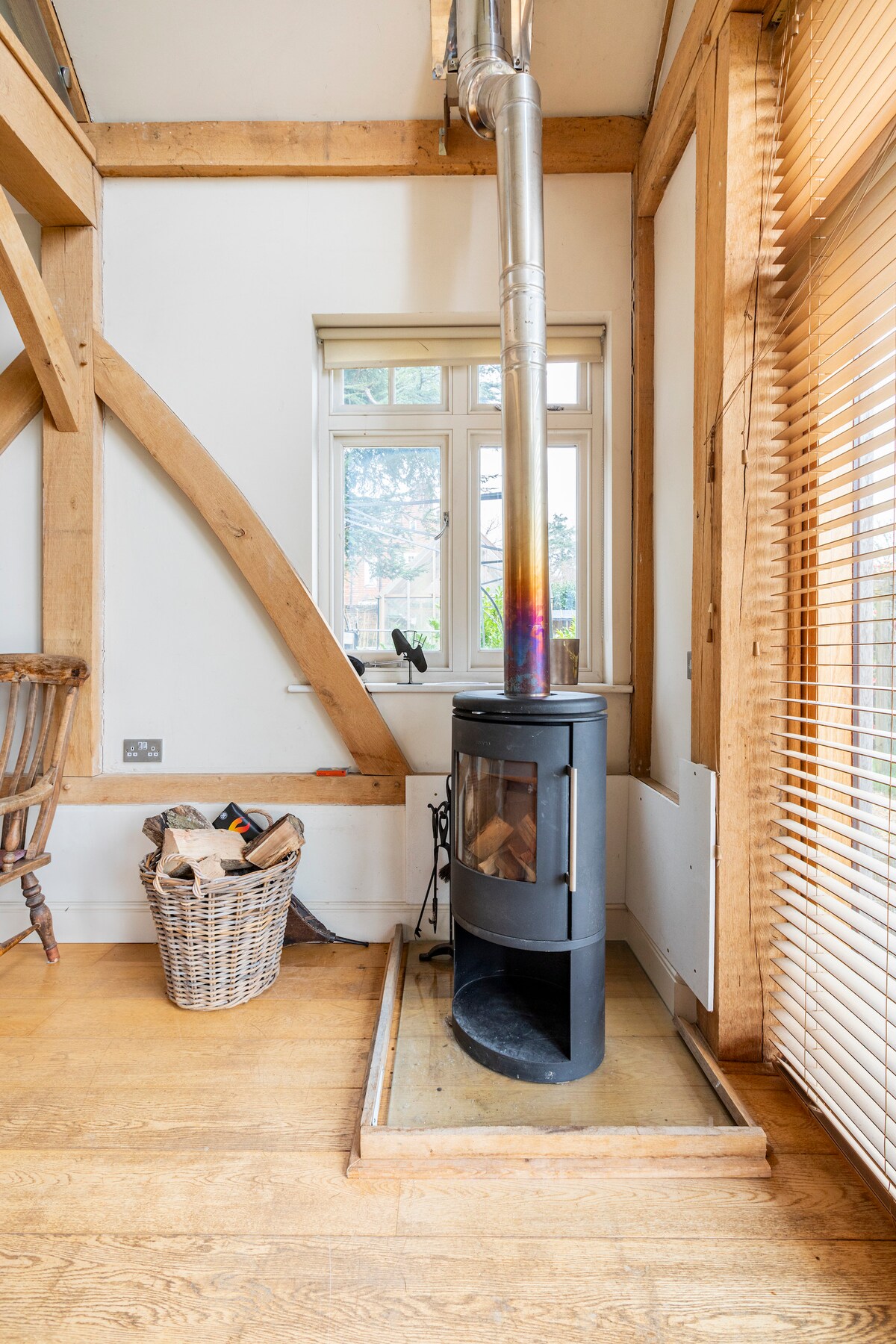 A black log burner is positioned beside a basket filled with firewood, set against a backdrop of wooden beams and a window. Natural light filters through partially closed blinds, highlighting the warm wooden flooring.