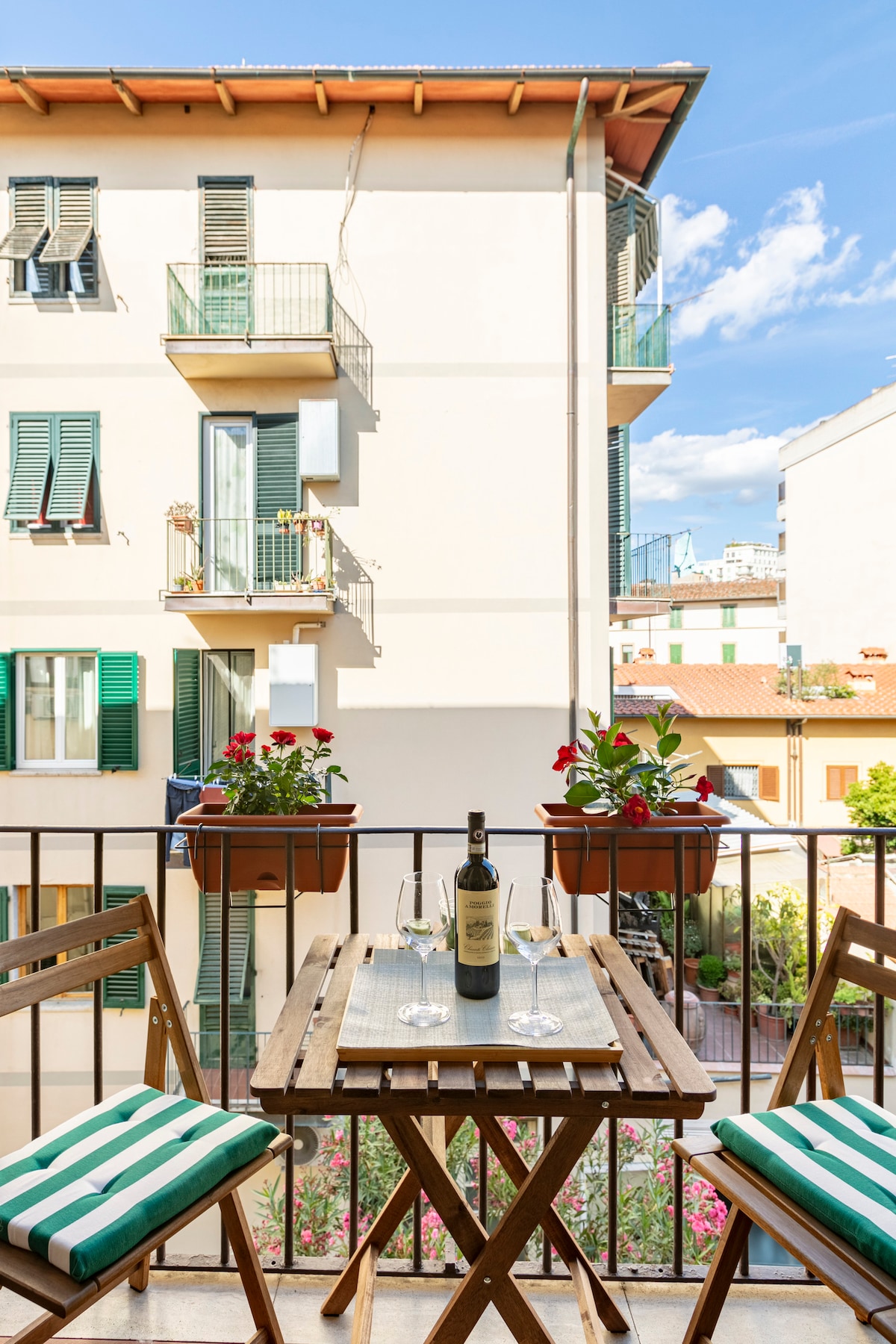A small balcony features a wooden table set with a bottle of wine and glasses, complemented by potted plants. The view includes buildings with green shutters and vibrant flowers below, enhancing the outdoor space's appeal.