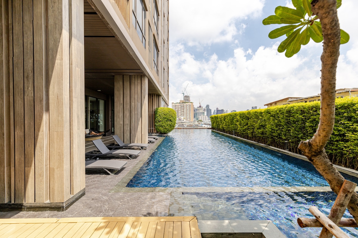 A serene swimming pool is framed by wooden decking and green hedges. Sun loungers are positioned alongside the pool, with the city skyline visible in the background. The area is designed for relaxation, offering a peaceful retreat in a central location.