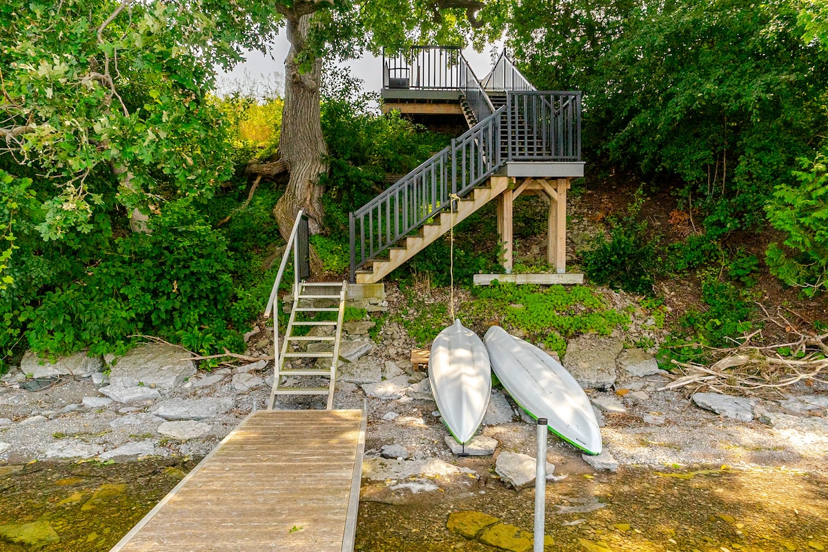 A wooden dock extends over a rocky shoreline, where two kayaks are secured alongside. A staircase leads up to a second-level platform nestled among trees, providing access to the suite above. Surrounding greenery enhances the natural setting by the lake.